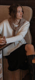 Woman sitting at a cafe table holding a cup, wearing a white sweater and black skirt.