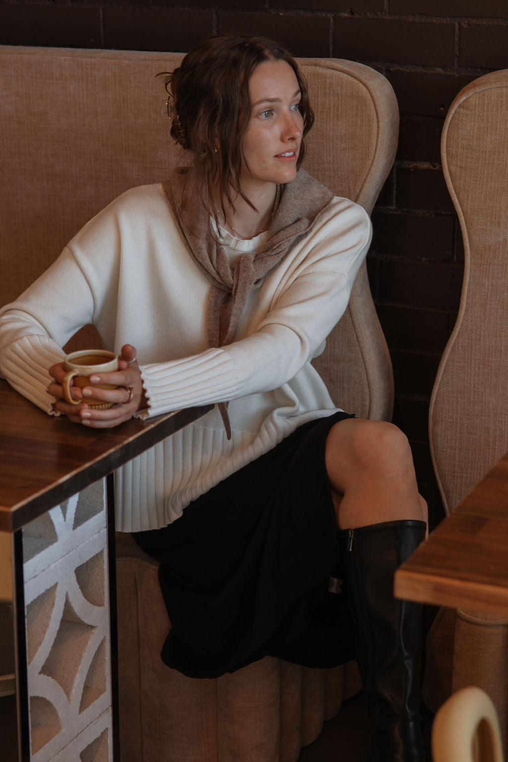 Woman sitting at a cafe table holding a cup, wearing a white sweater and black skirt.