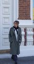 Person wearing a long coat standing in front of a building with decorative white columns and a brick wall.