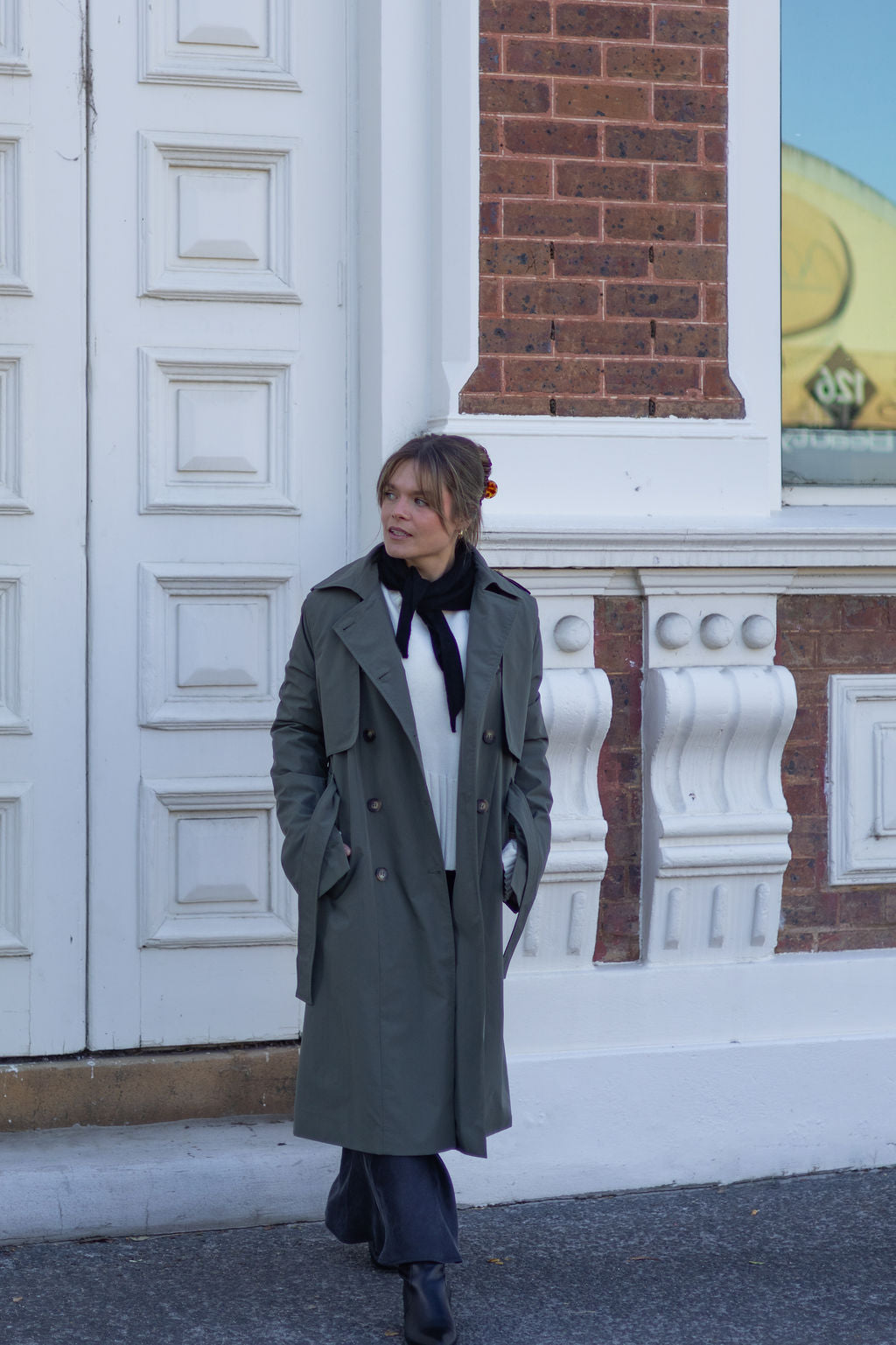 Person wearing a long coat standing in front of a building with decorative white columns and a brick wall.