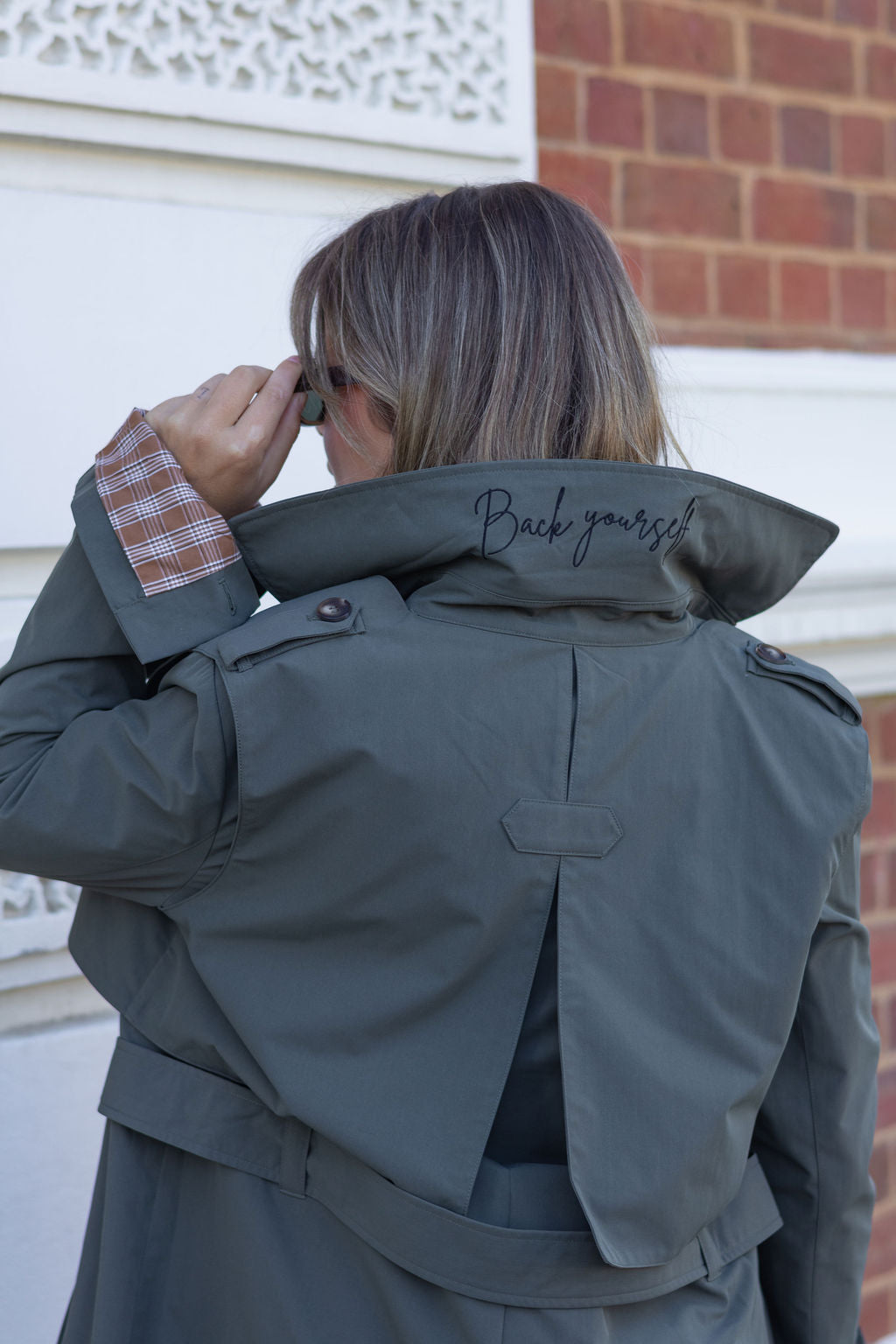 Person wearing a gray jacket with 'Back yourself!' text on the back, standing in front of a building.