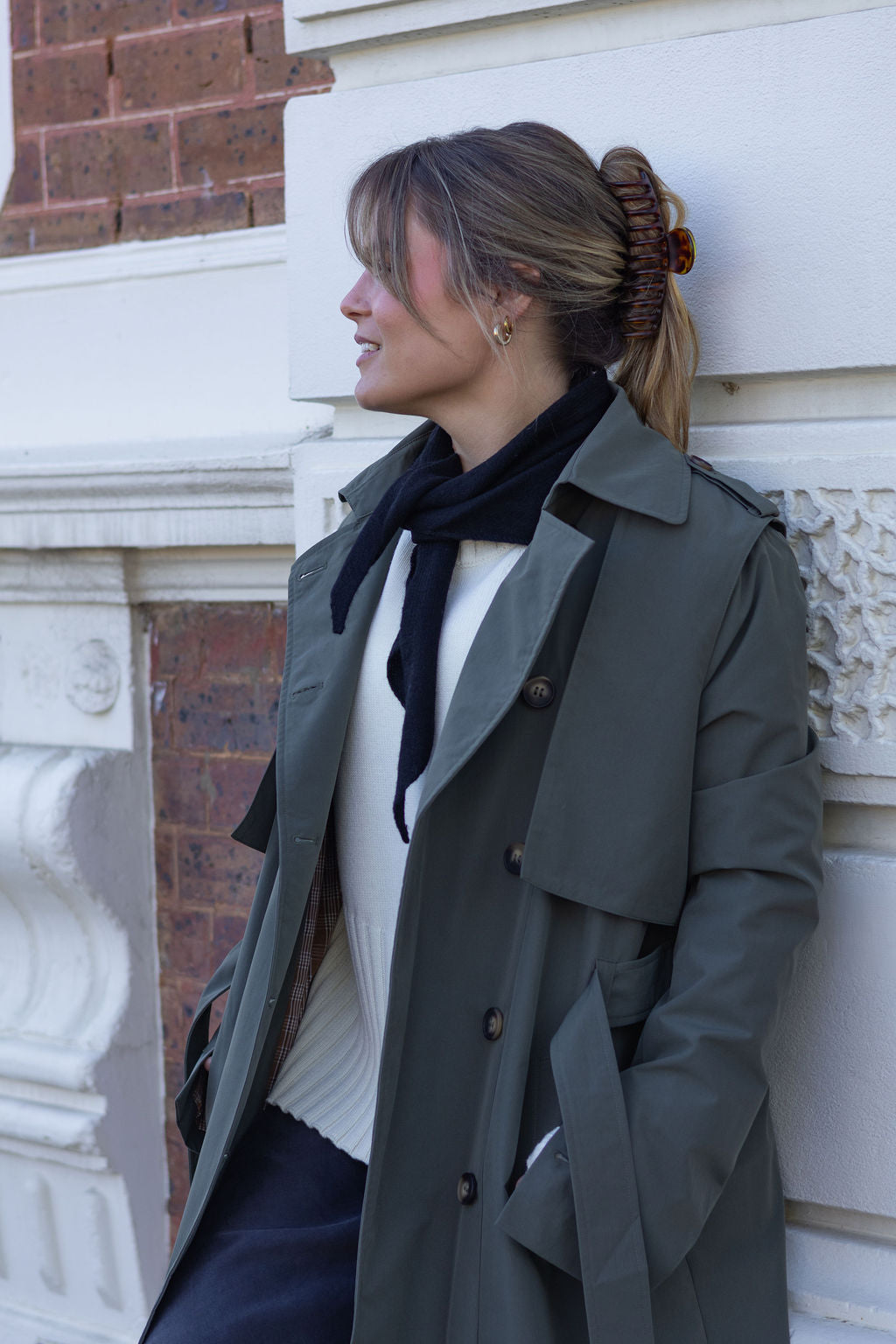 Woman wearing a green coat and black scarf leaning against a building.