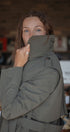 Person wearing a green coat with a brand logo, standing in a blurred indoor setting.