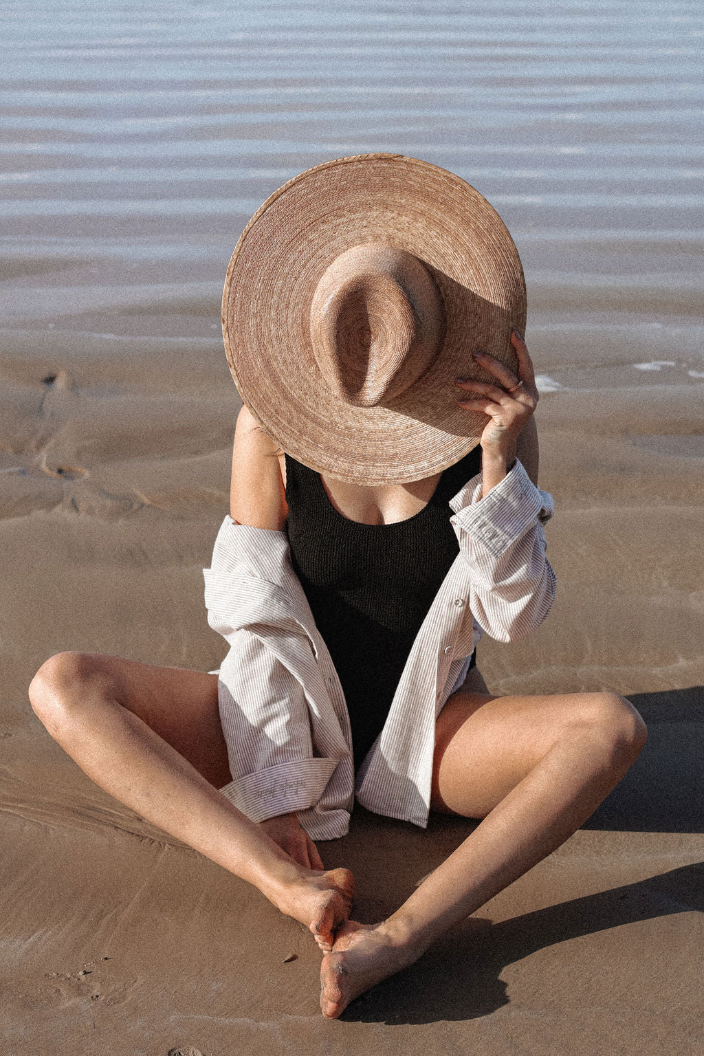 Person sitting on sand wearing a wide-brimmed hat and light shirt.