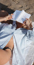 Person lying on a beach reading a book with a white shirt and black bikini bottom.