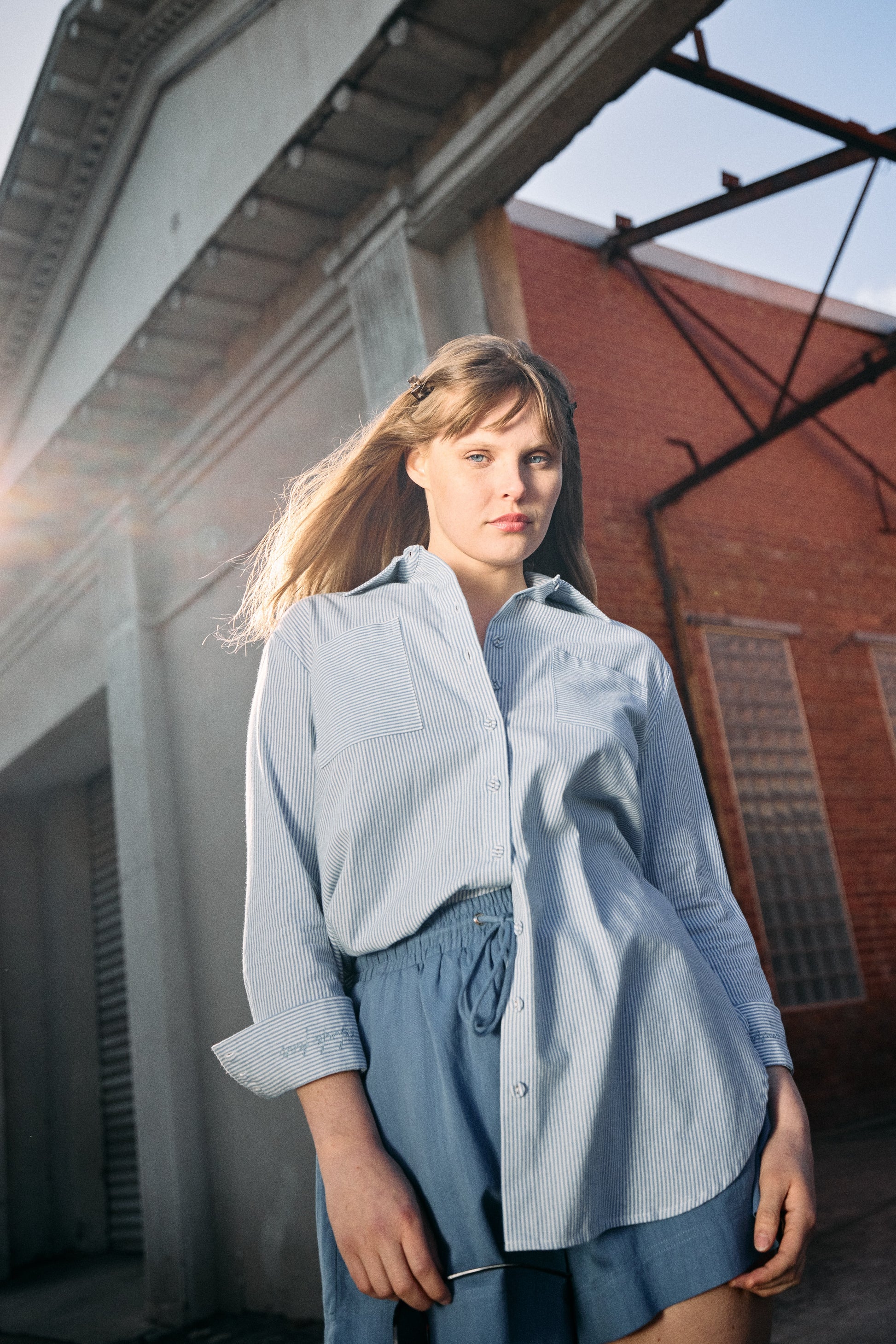 Woman wearing a light blue shirt and shorts standing in front of a brick building.