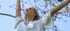 Woman in a white dress with arms raised against a blue sky with tree branches.