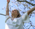 Woman in a white dress with arms raised against a blue sky with tree branches.