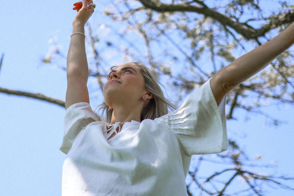 Woman in a white dress with arms raised against a blue sky with tree branches.