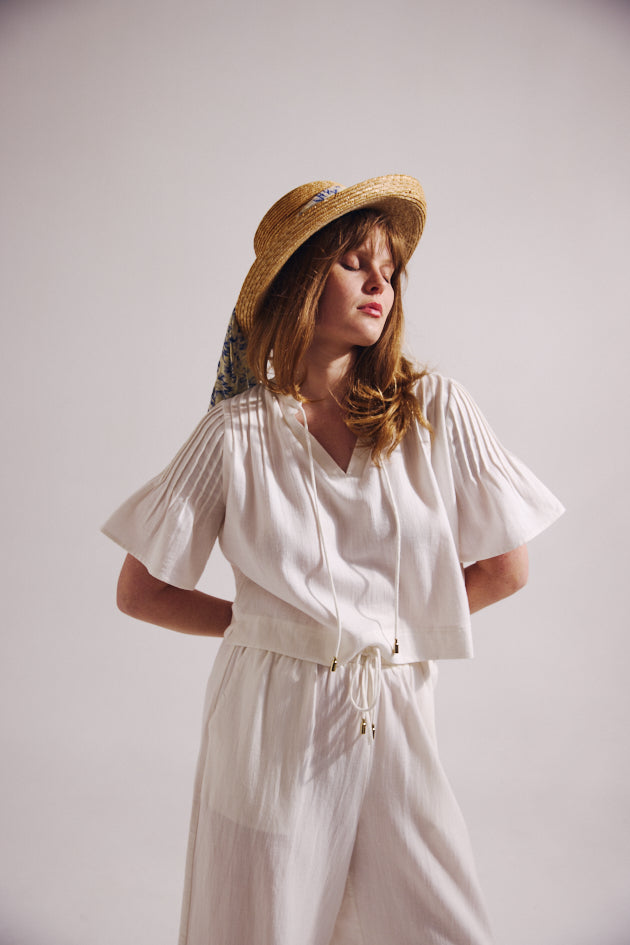 Woman wearing a white blouse and white pants with a straw hat on a plain background