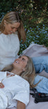 Two women sitting on a blanket in a garden with flowers and a basket.