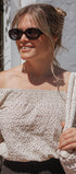 A woman wearing a cream off-the-shoulder top with a small brown floral pattern, styled with black sunglasses and holding a chunky knit cream bag against a white stucco wall background.