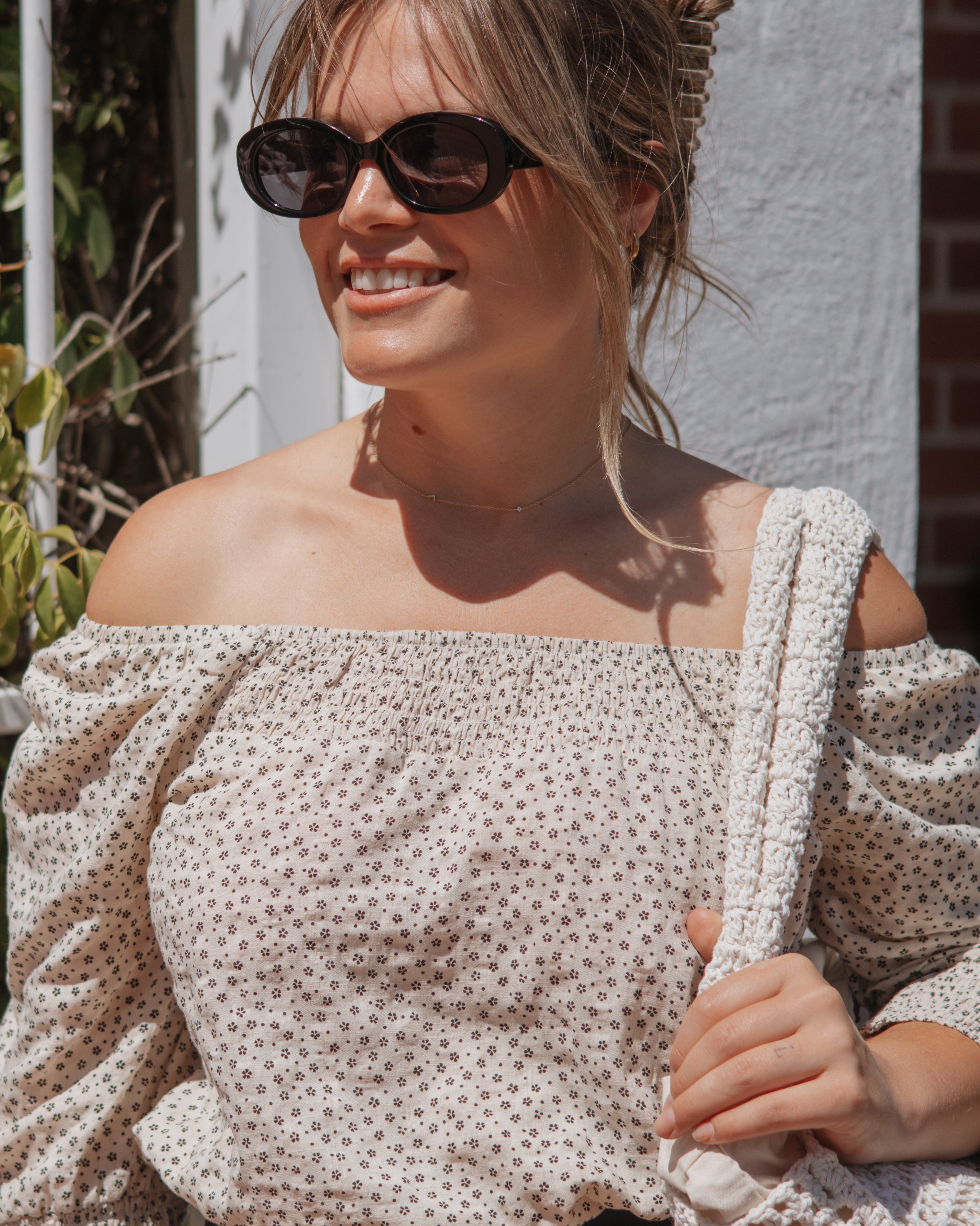 A woman wearing a cream off-the-shoulder top with a small brown floral pattern, styled with black sunglasses and holding a chunky knit cream bag against a white stucco wall background.