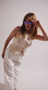 Woman wearing a beige sleeveless top and white pants, adjusting sunglasses against a plain background