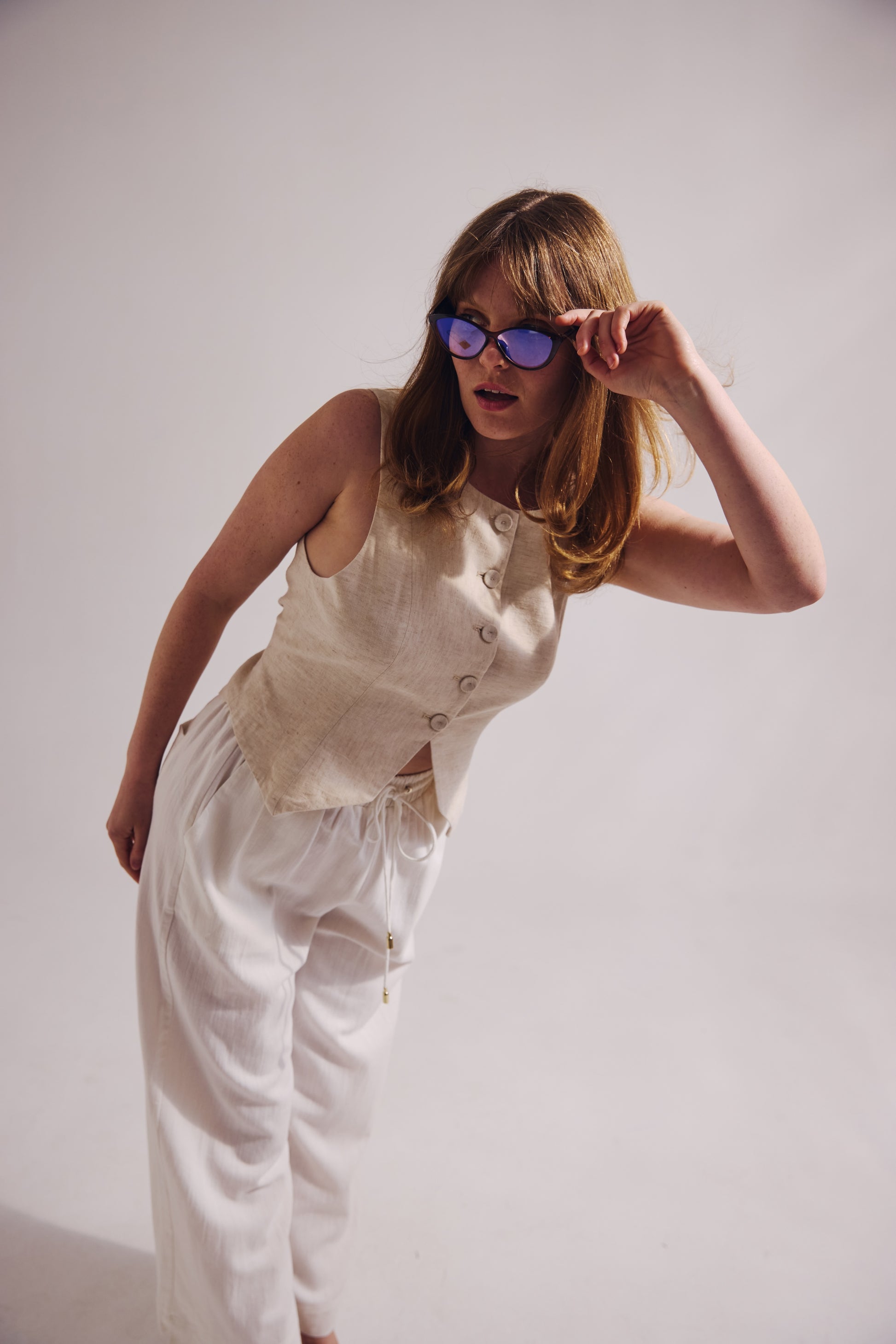 Woman wearing a beige sleeveless top and white pants, adjusting sunglasses against a plain background