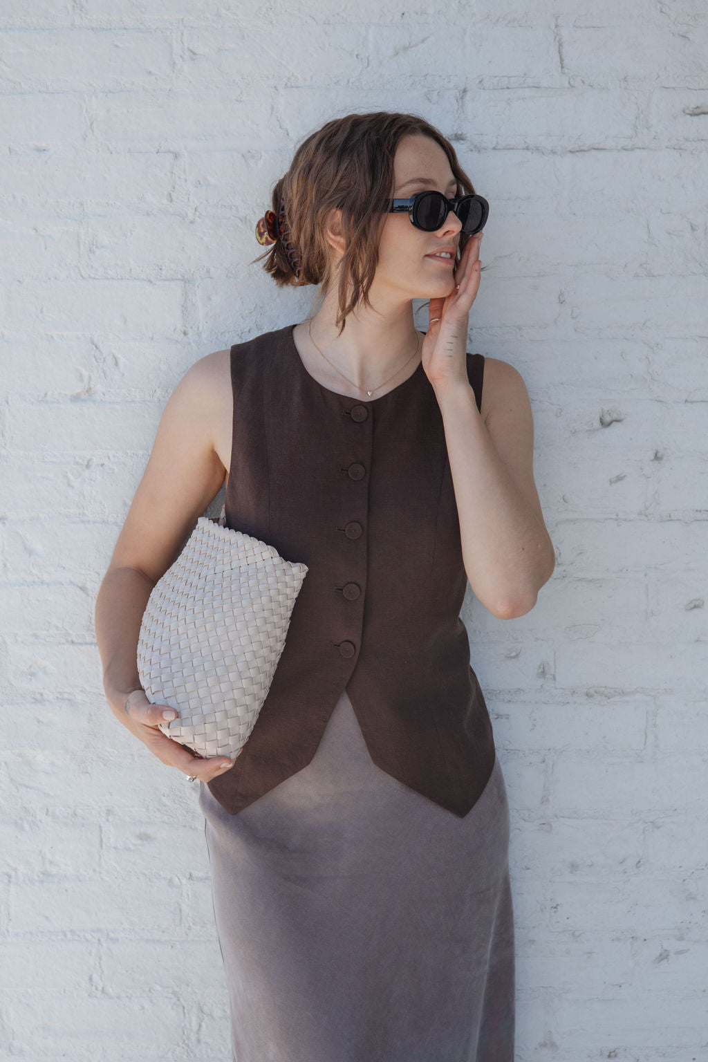 Woman wearing a brown vest over a gray dress against a white brick wall.