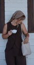 Woman in brown outfit holding a cup and a woven bag against a white wall with wooden frames.