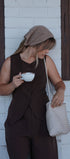 Woman in brown outfit holding a cup and a woven bag against a white wall with wooden frames.