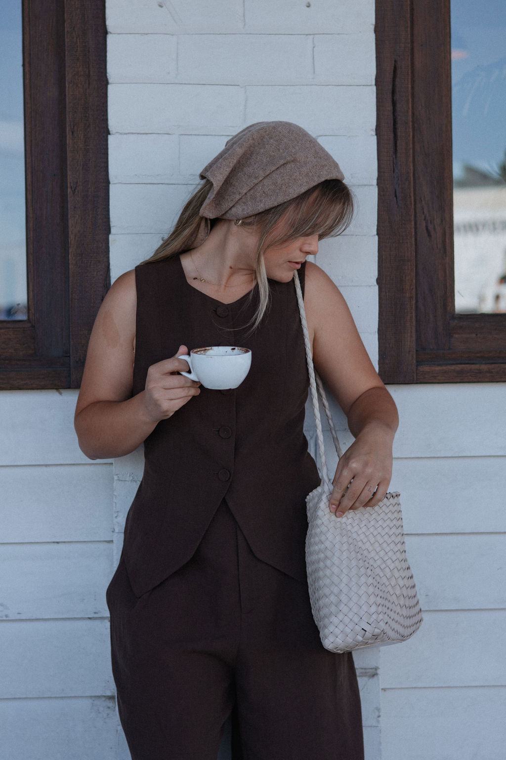 Woman in brown outfit holding a cup and a woven bag against a white wall with wooden frames.