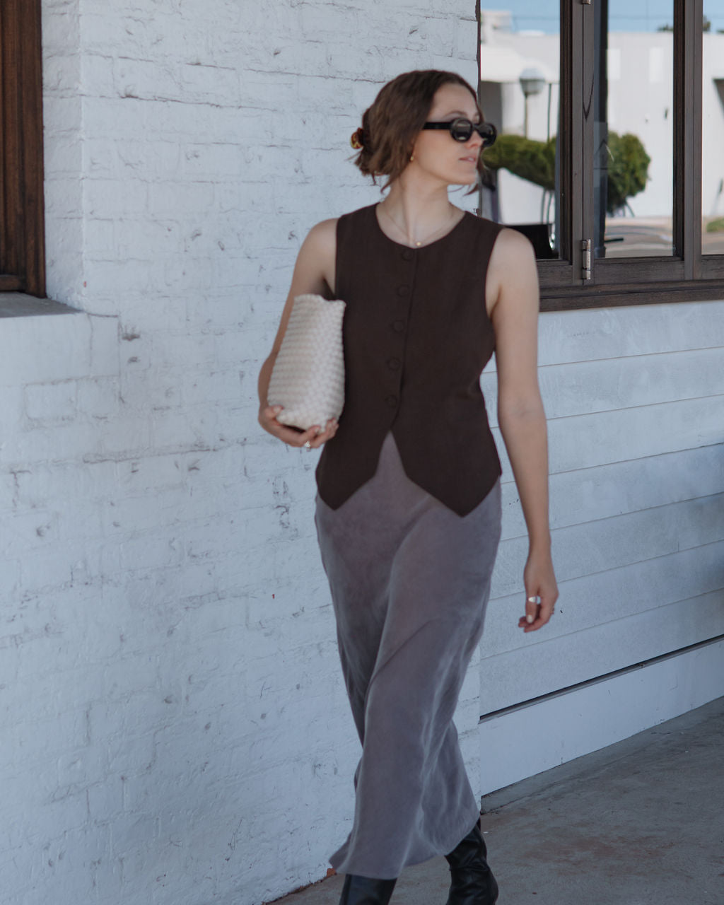 Woman in a sleeveless brown top and gray pants standing against a white wall.