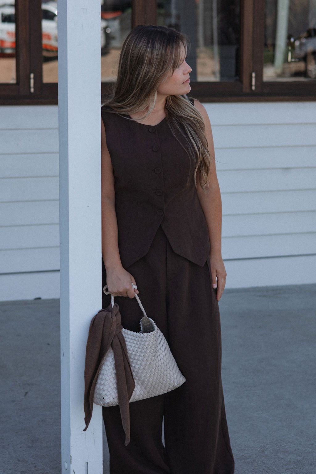 Woman in a brown outfit holding a white handbag against a building background