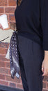 Person wearing a black outfit with a patterned scarf, holding a white cup against a brick wall.