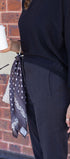 Person wearing a black outfit with a patterned scarf, holding a white cup against a brick wall.