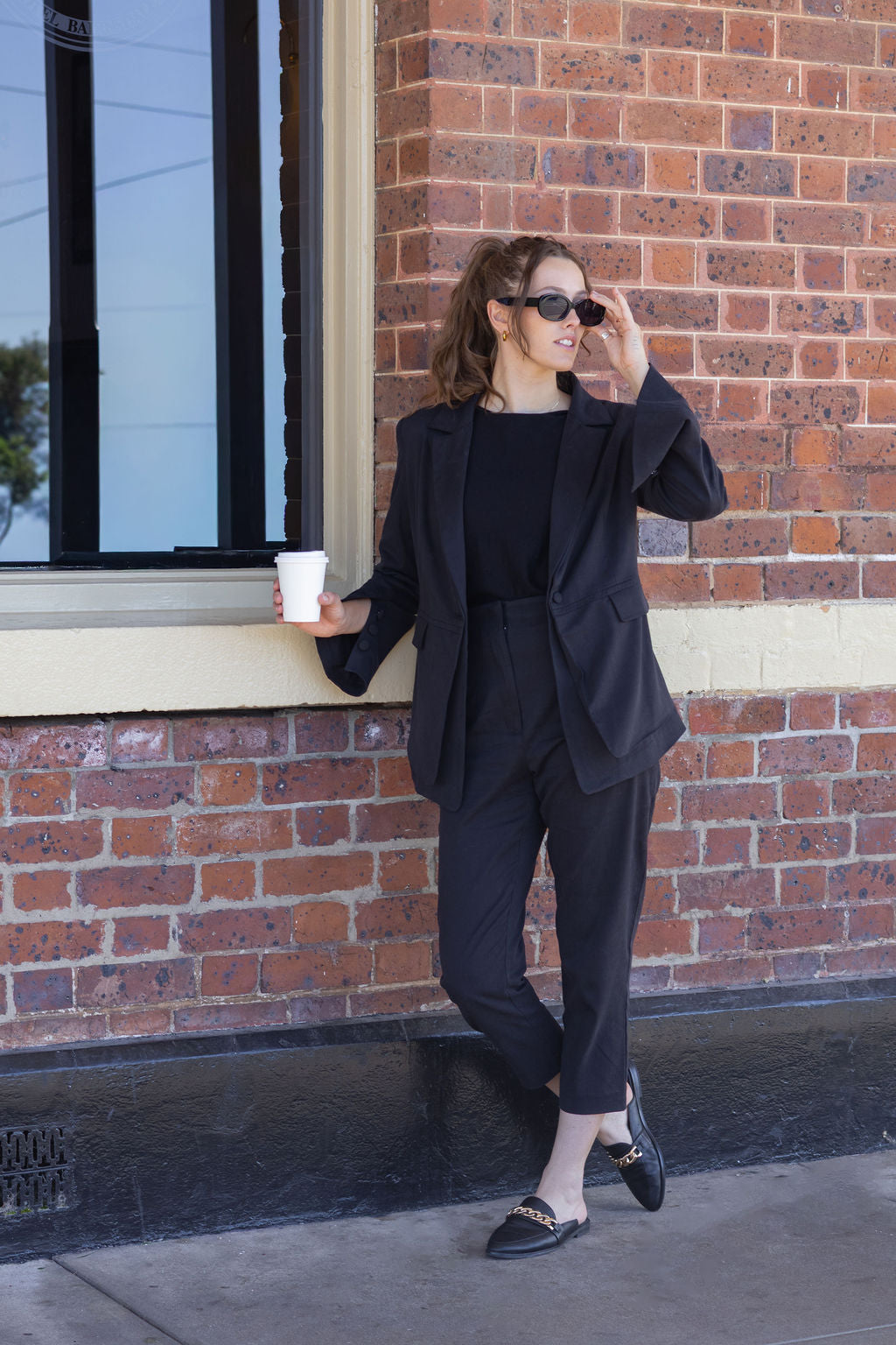 Woman in a black suit holding a coffee cup against a brick wall.