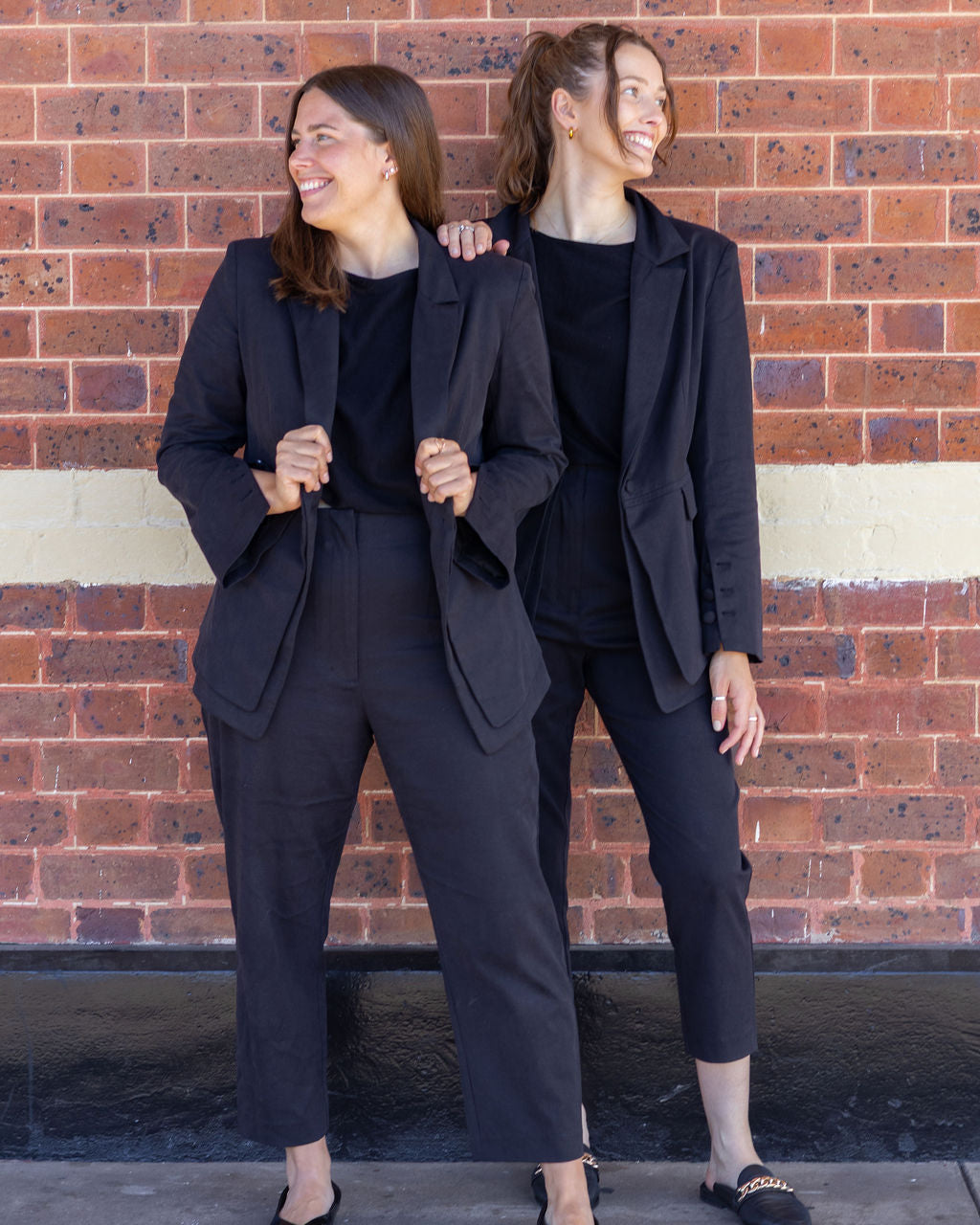 Two women in matching black suits standing against a brick wall.