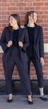 Two women in matching black suits standing against a brick wall.