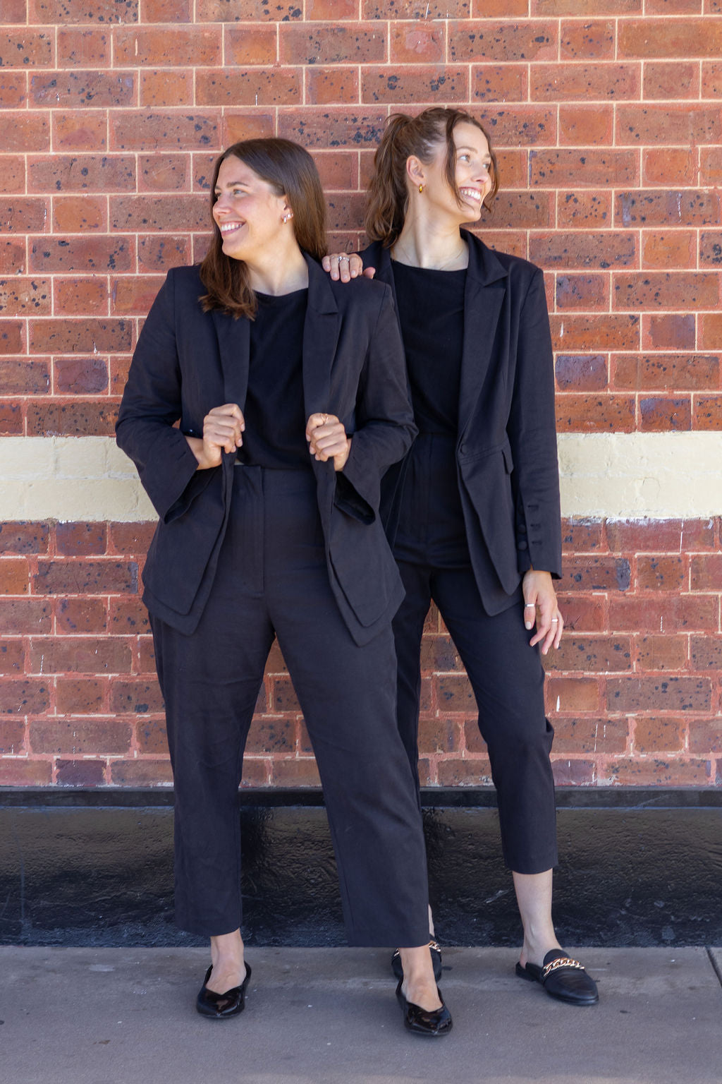 Two women in matching black suits standing against a brick wall.