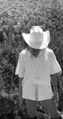 Person wearing a white cowboy hat and outfit standing in a field of tall grass.