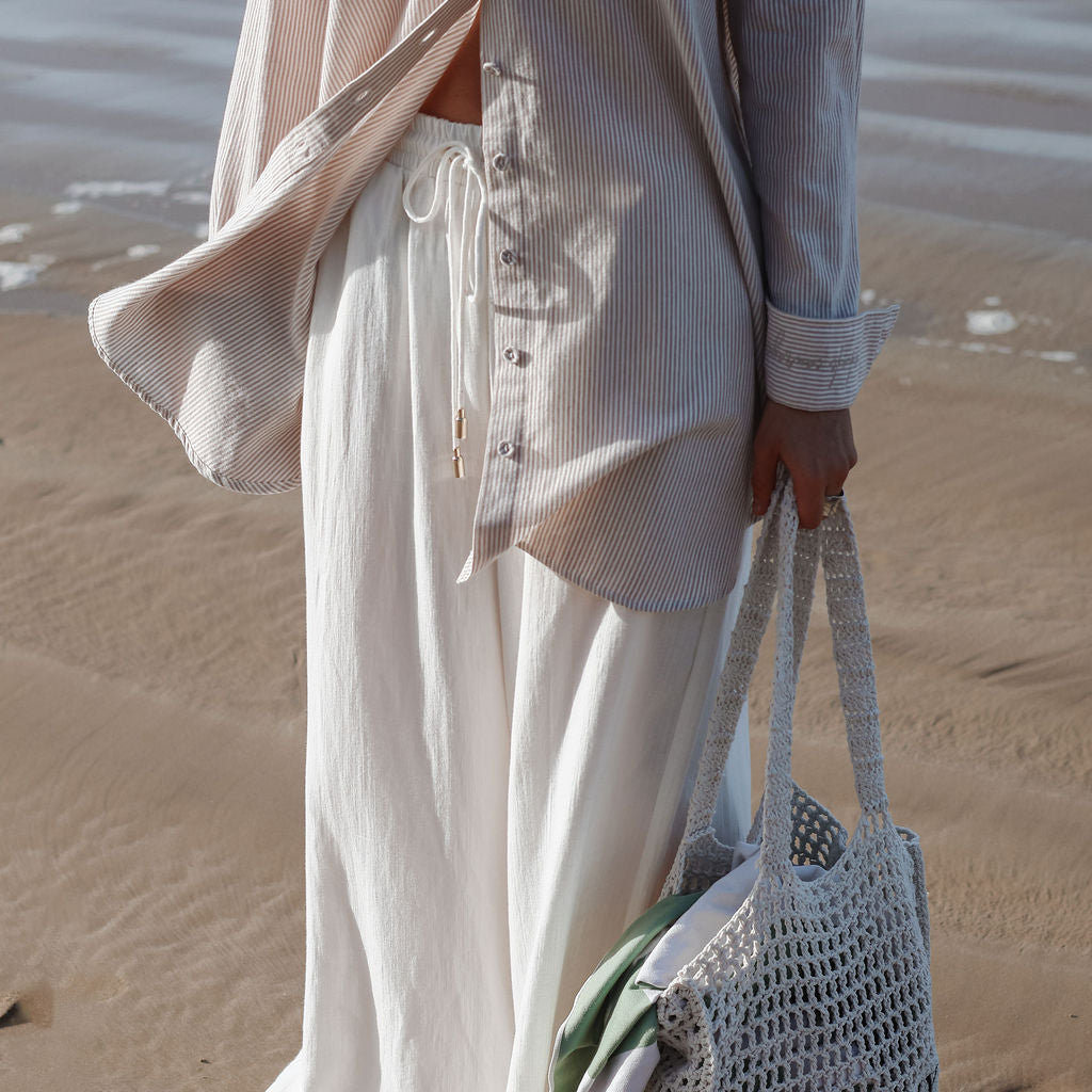 Person on a beach wearing a light outfit with a woven bag.