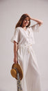 Woman in a white dress holding a straw hat against a plain background