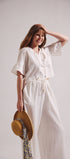 Woman in a white dress holding a straw hat against a plain background