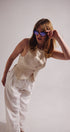 Woman wearing a beige top and white pants with sunglasses on a plain background