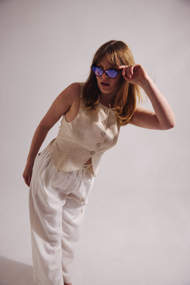 Woman wearing a beige top and white pants with sunglasses on a plain background