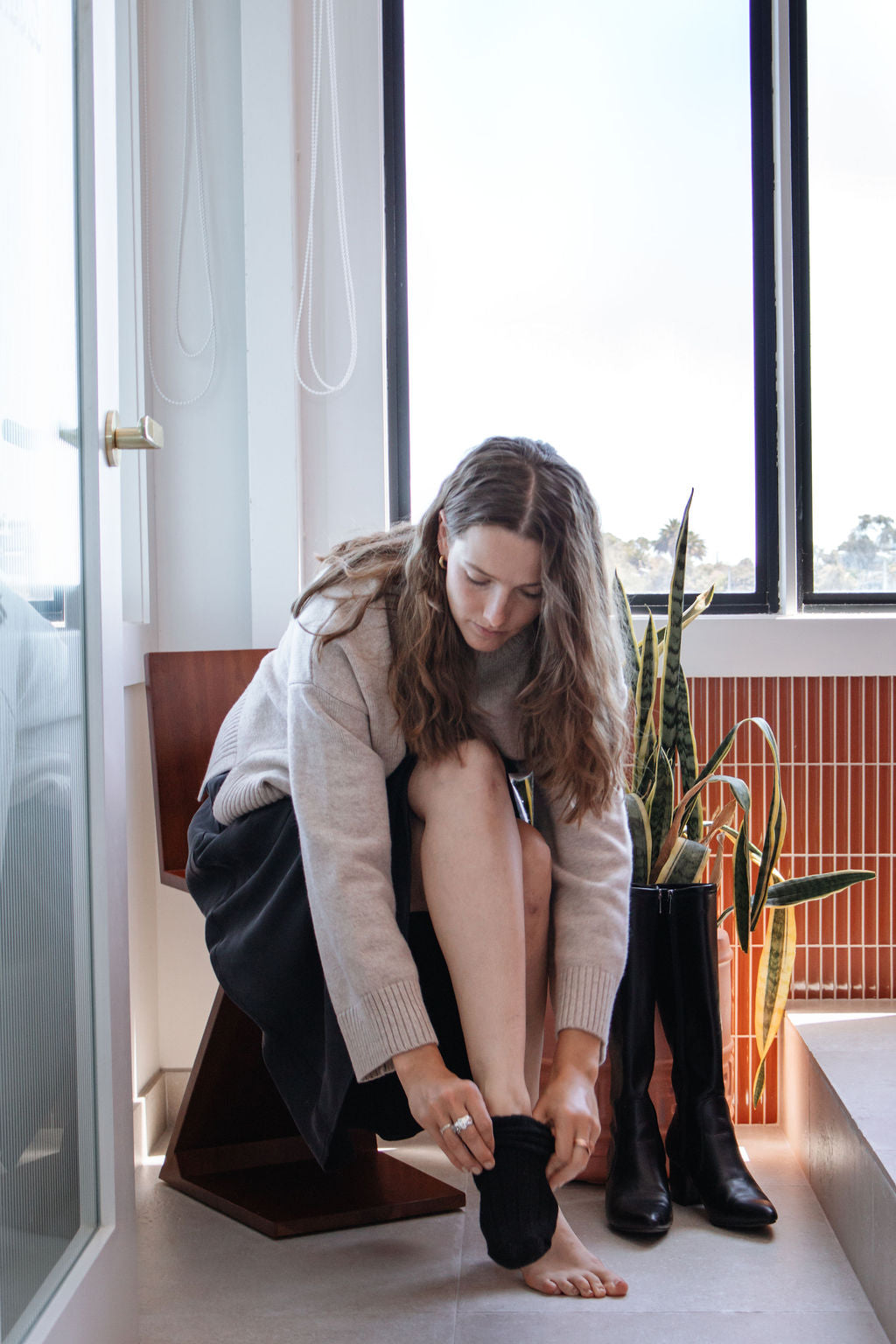 Woman sitting on a chair by a window, putting on black boots.