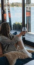 Woman sitting on a couch by large windows with a view of water, holding a glass of red wine.