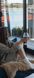 Woman sitting on a couch by large windows with a view of water, holding a glass of red wine.
