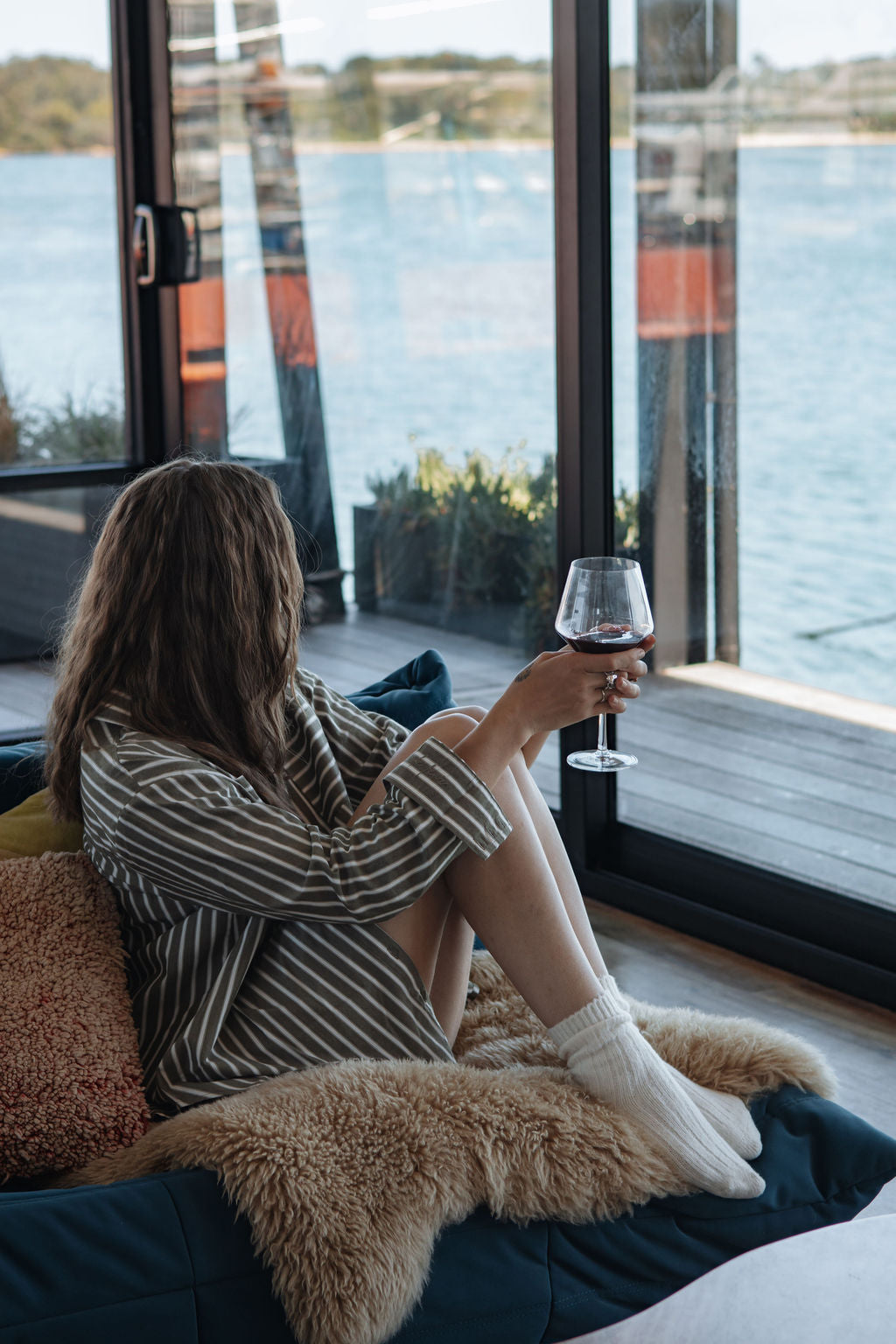 Woman sitting on a couch by large windows with a view of water, holding a glass of red wine.