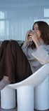 Woman sitting on a modern white chair by a large window, holding a mug.