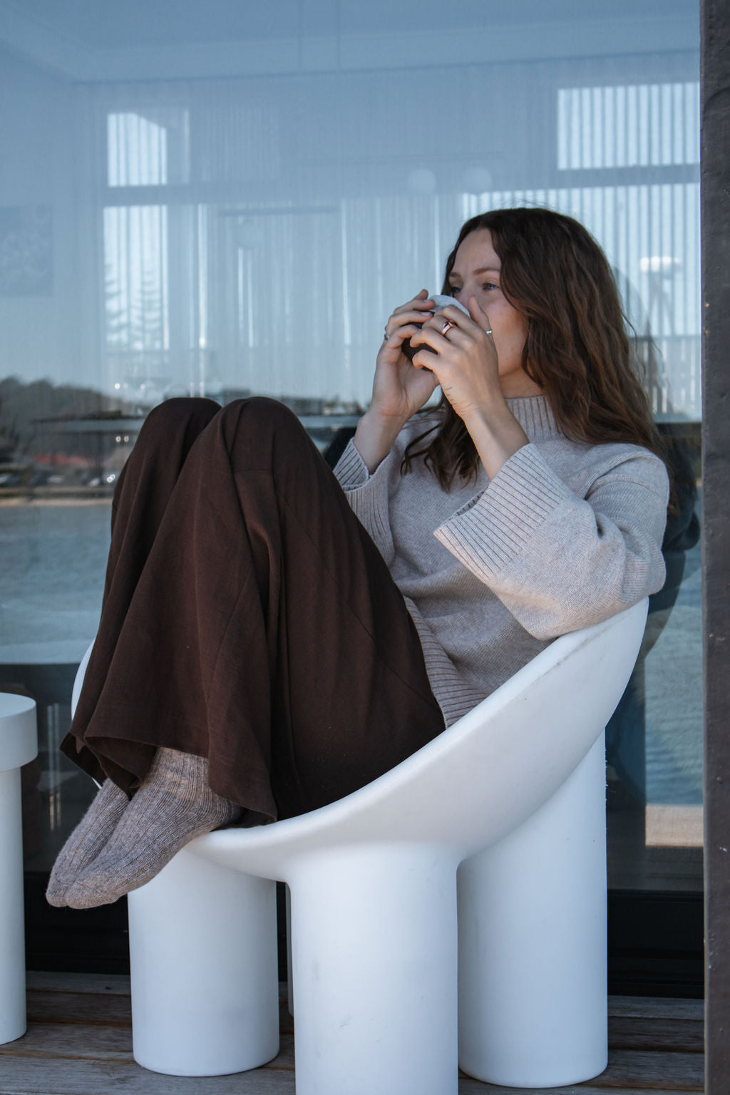 Woman sitting on a modern white chair by a large window, holding a mug.