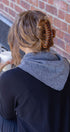 Person with a hair curler in their hair, wearing triangle scarf, sitting against a brick wall holding a cup.
