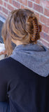 Person with a hair curler in their hair, wearing triangle scarf, sitting against a brick wall holding a cup.