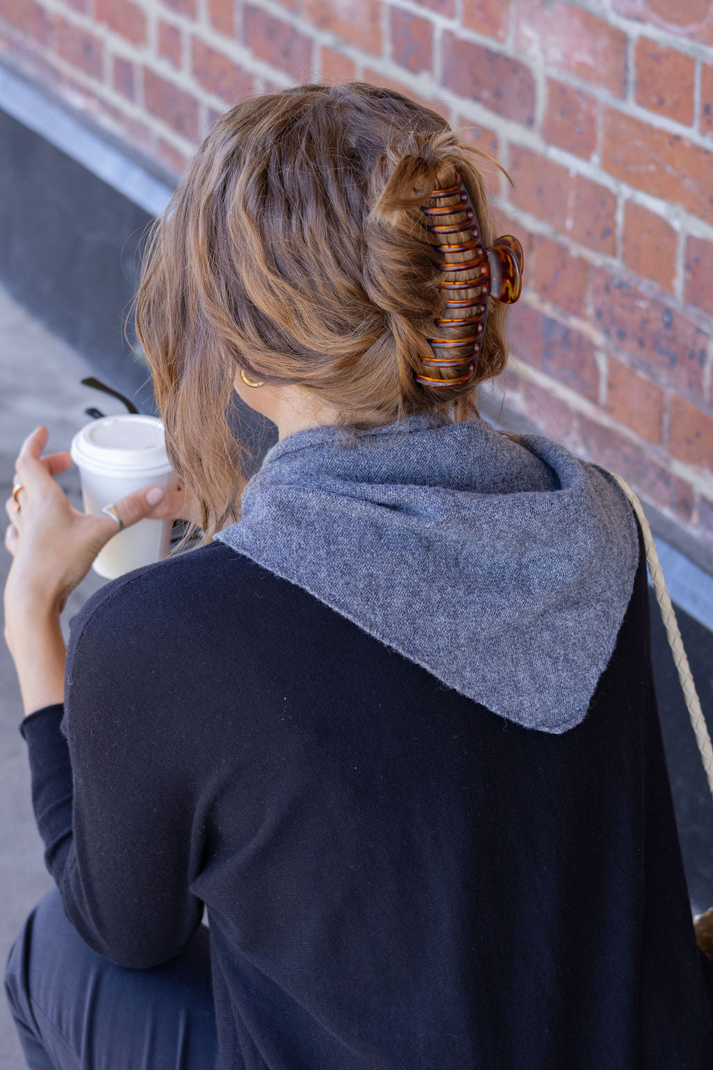 Person with a hair curler in their hair, wearing triangle scarf, sitting against a brick wall holding a cup.