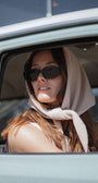 Woman wearing sunglasses and a headscarf, sitting inside a vehicle.