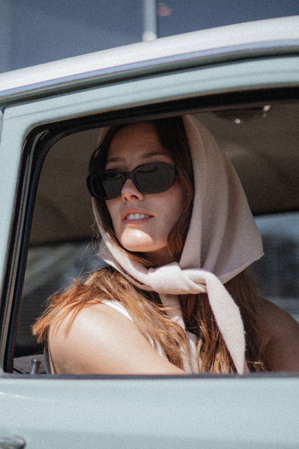Woman wearing sunglasses and a headscarf, sitting inside a vehicle.